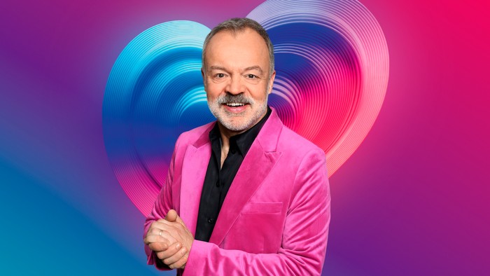 565007,TITLE:Eurovision Song Contest 2026 Graham Norton wearing a black shirt, crushed pink velvet blazer stood smiling ahead with his hands placed together.