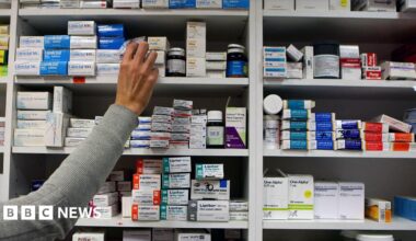 A white shelf full of medication boxes and a hand reaching up to get a box