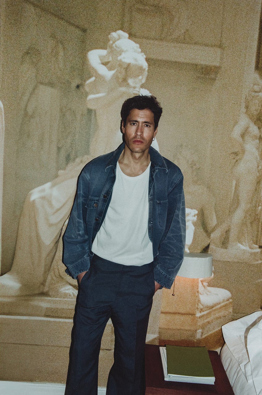 Man in a denim jacket and trousers standing in front of a classical sculpture backdrop.
