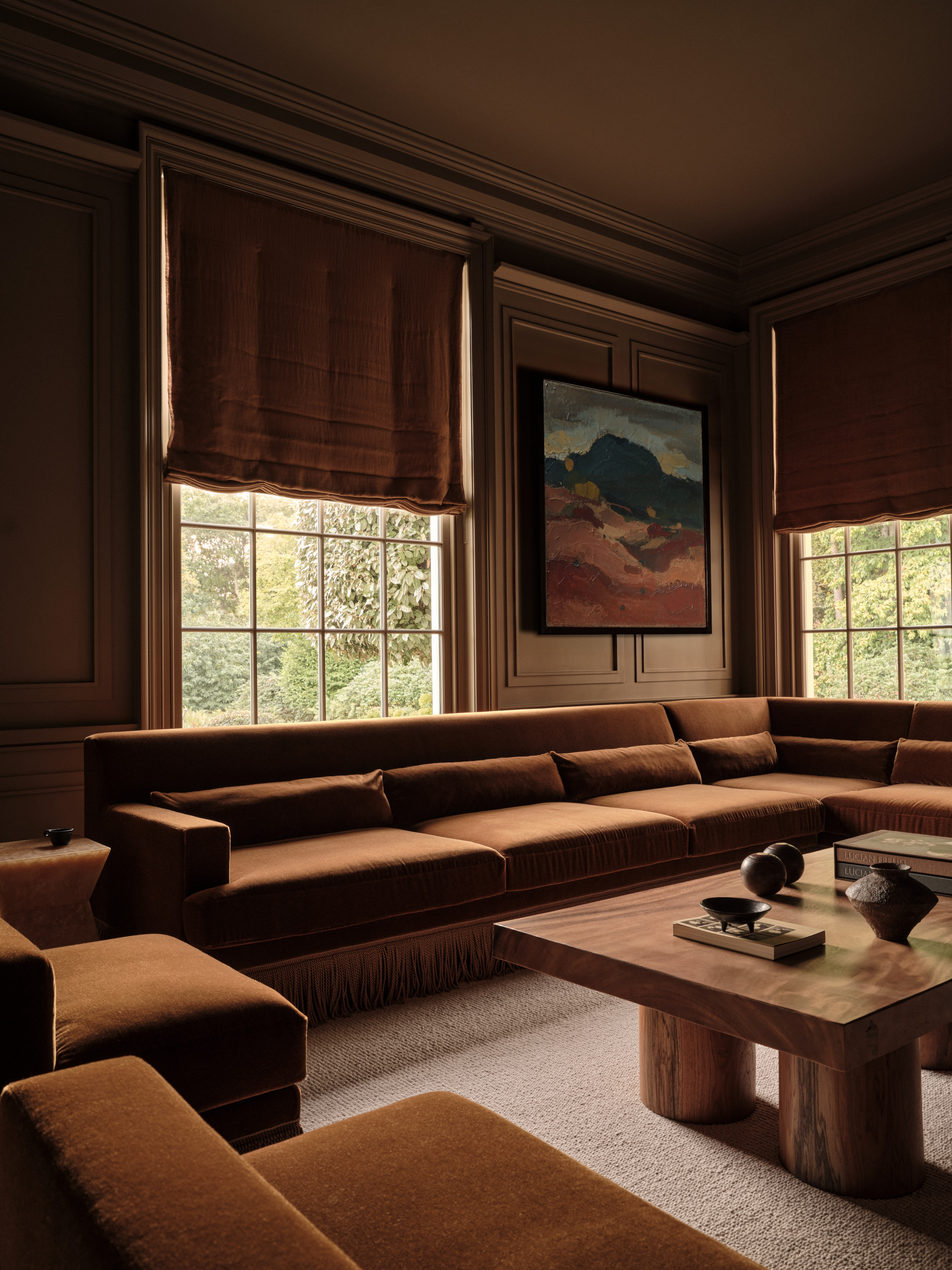 Color-drenched burnt orange snug with fringed corner sofa, Roman blinds, chunky wood coffee table and landscape artwork