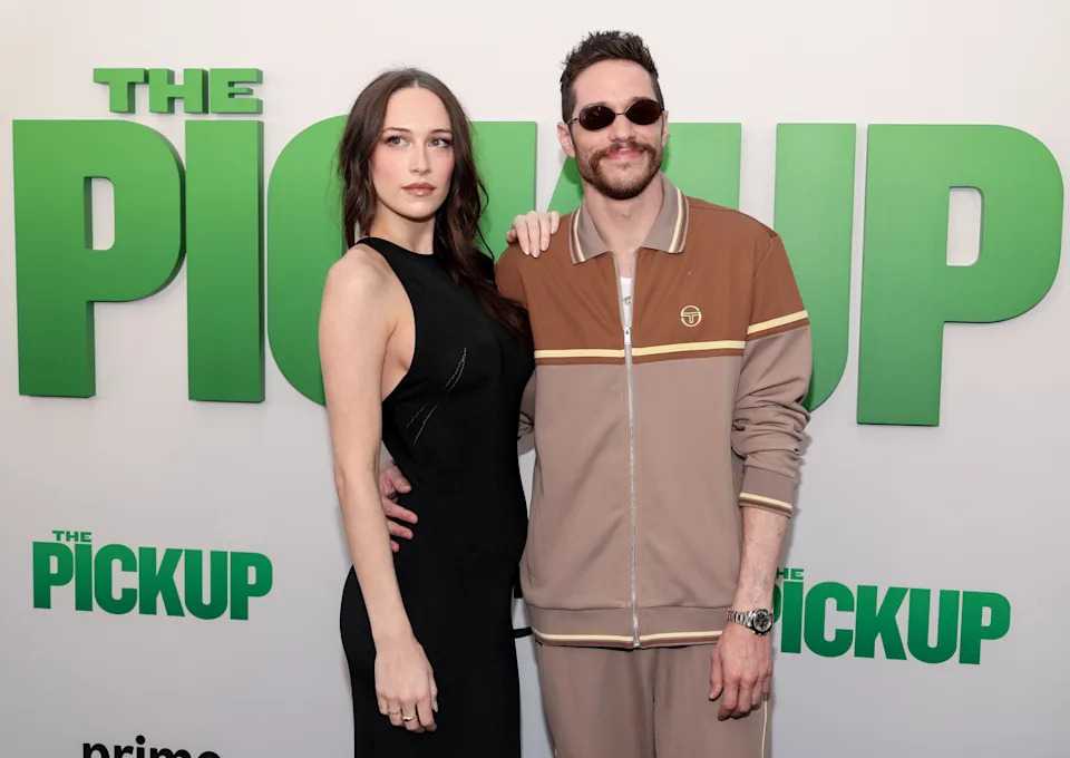 Two individuals pose at a "The Pickup" event. The person on the left wears a sleek black dress, and the person on the right is in a casual brown tracksuit