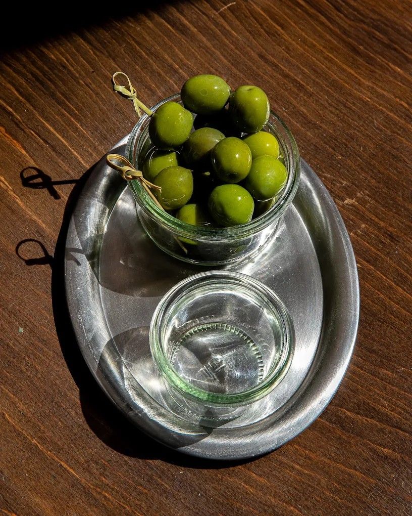 A chrome serving platter, topped with glass jars filled with olives, and placed atop a wooden table.