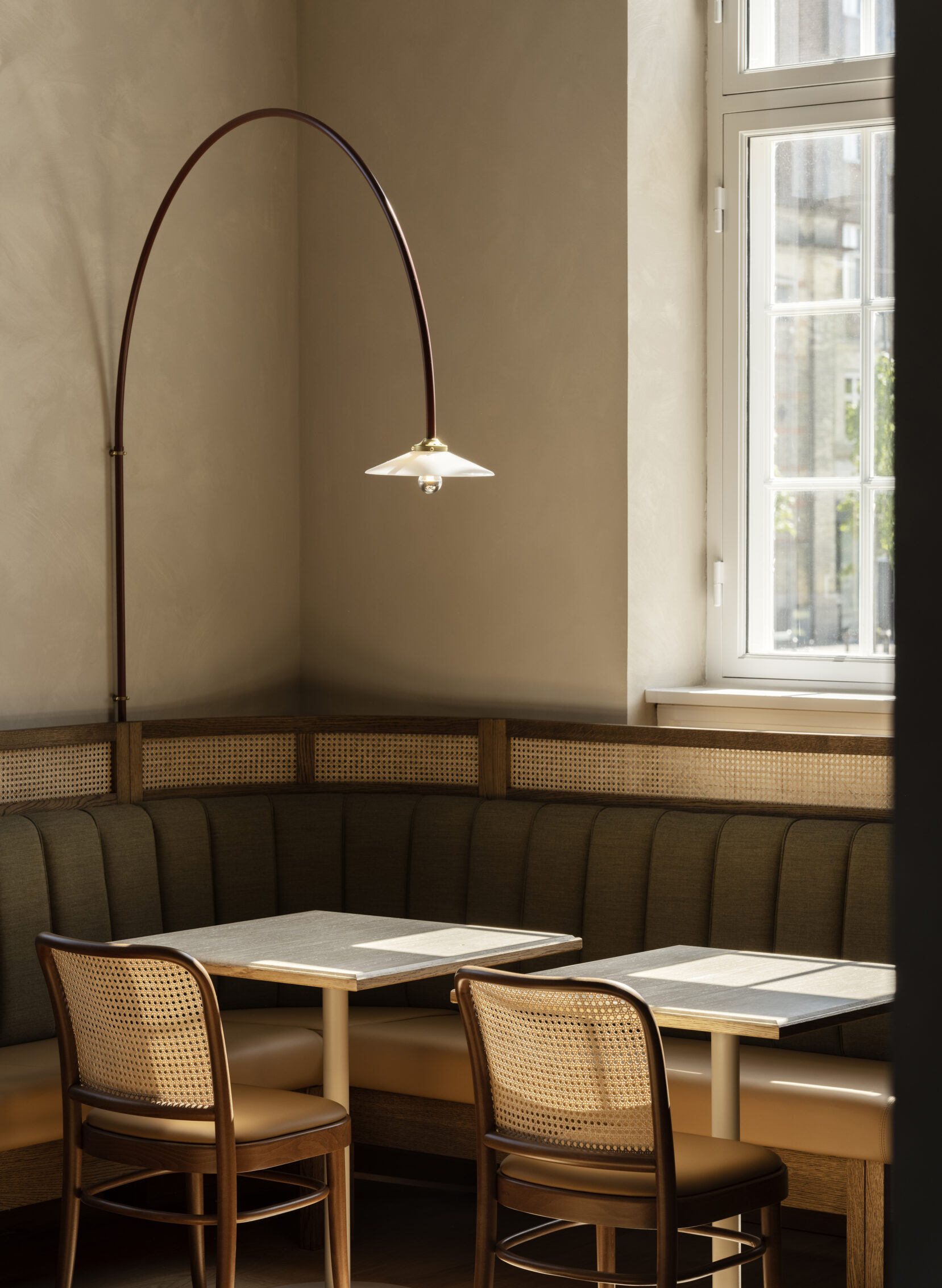A sun-lit dining room features retro-inspired, bistro-like interiors, with olive green banquette seating, wooden chairs with rattan, and marble tables, as well as arched Art Deco lighting.