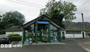 Google Maps image of Coniston Visitor Information Centre. It has a slanted roof, like an upturned V, and glass frontage. Trees and hills can be seen in the background.
