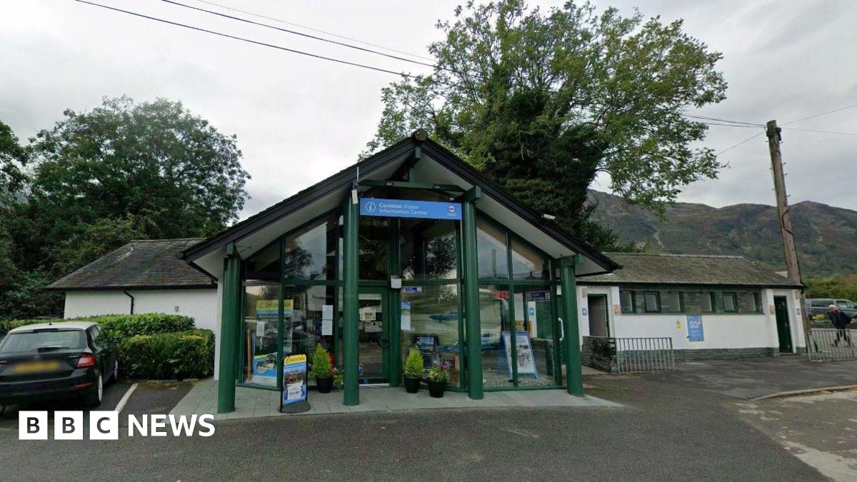 Google Maps image of Coniston Visitor Information Centre. It has a slanted roof, like an upturned V, and glass frontage. Trees and hills can be seen in the background.
