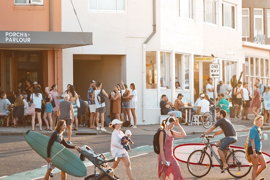 People walk with surfboards and ride bikes across a road with people standing and sitting at cafes.