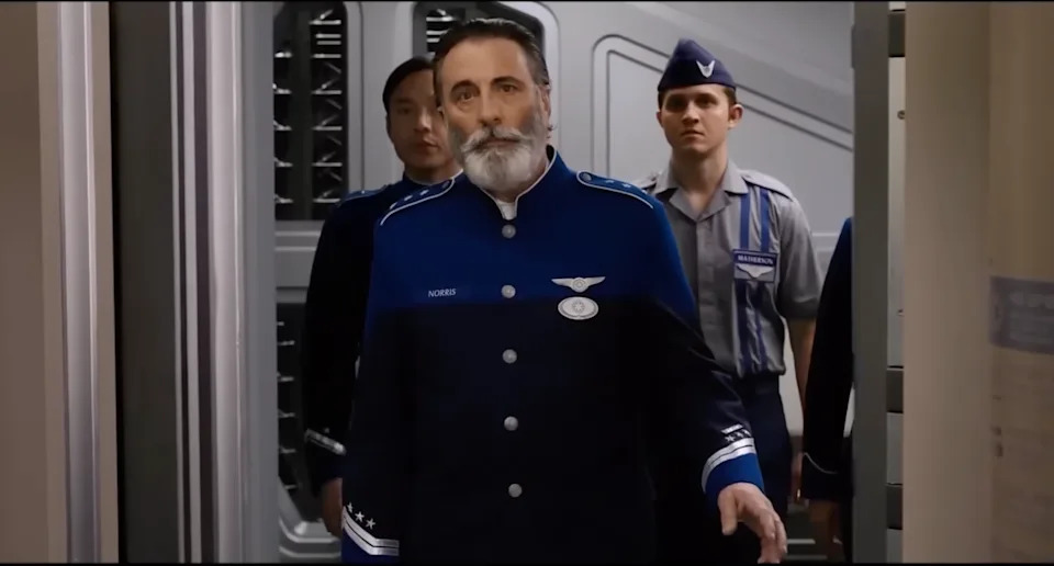 A man in a blue military uniform with a beard walks in a futuristic setting, followed by others in similar attire