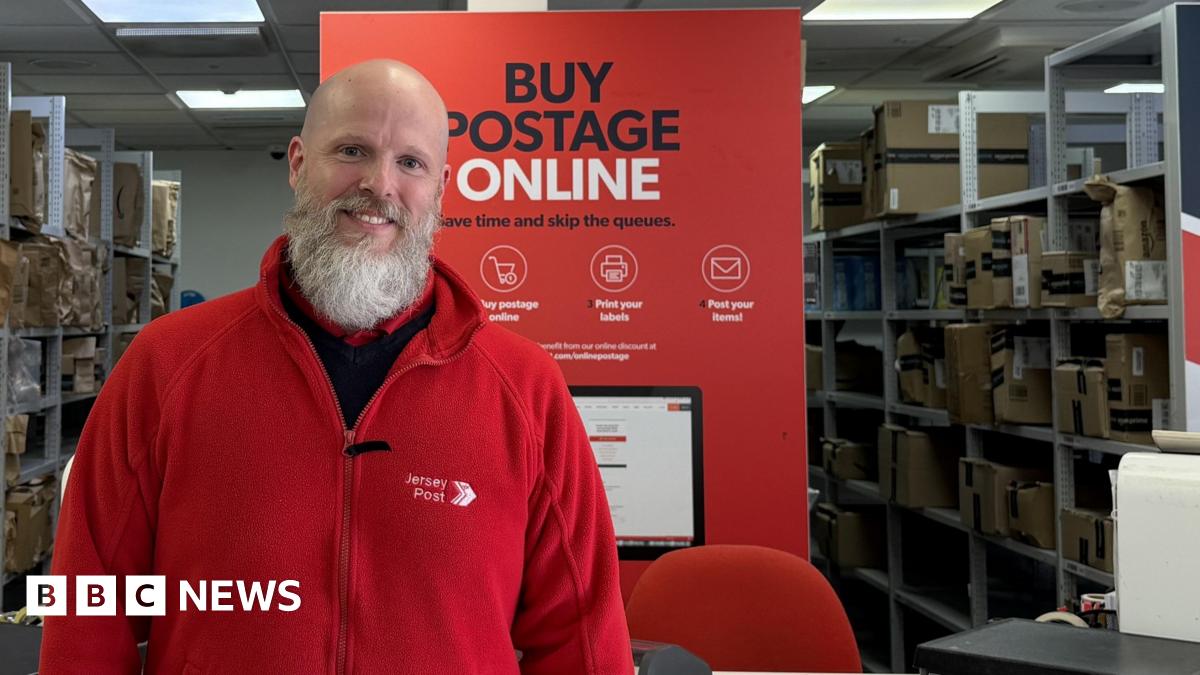 Chris wearing his red Jersey Post fleece jacket standing in front of the counter at Jersey Post headquarters. A sign behind him says "Buy postage online: Save time and skip the queues."
