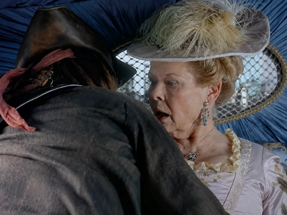 A person in a vintage outfit and hat looks surprised, interacting with someone in an old-fashioned jacket, possibly from a historical film scene
