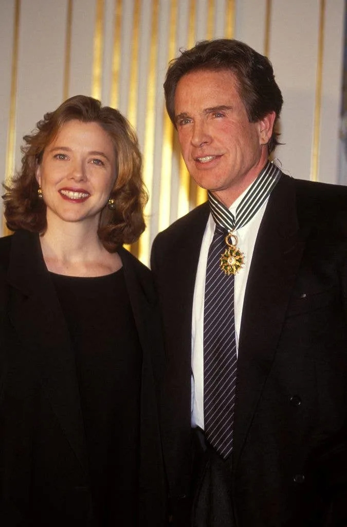 Warren Beatty and Annette Bening stand together in Paris. Annette wears a black top and jacket next to husband Warren who wears a white shirt, dark tie and black jacket with a medal around his neck.