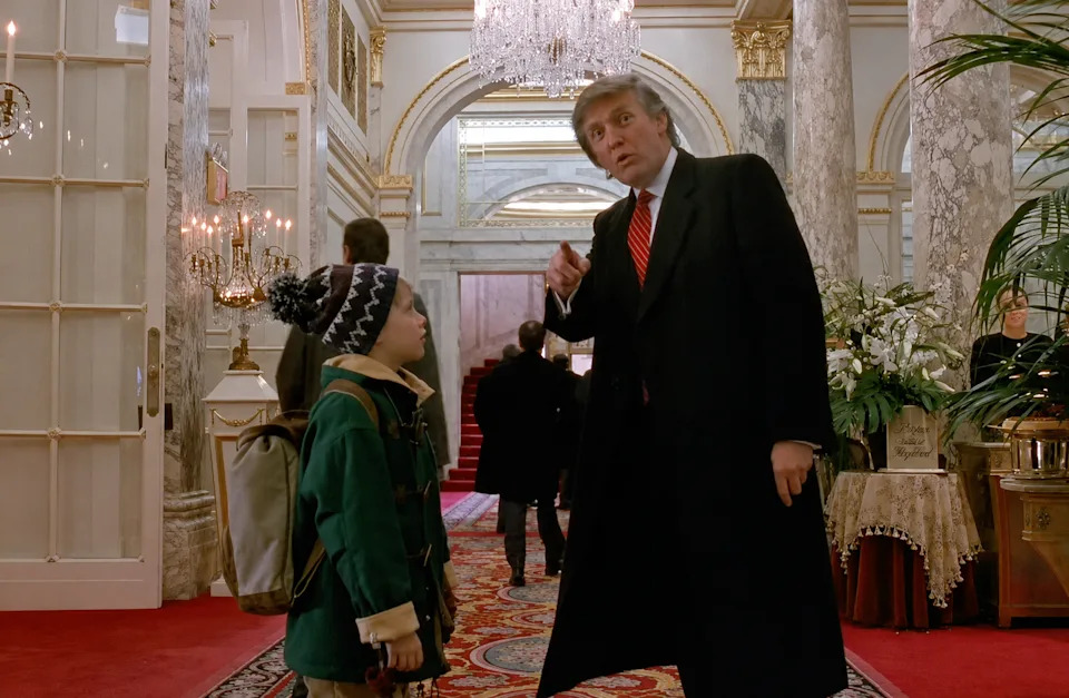 A young boy in a winter coat and hat stands in an elegant, ornate lobby talking to a man in a suit and tie pointing towards the hallway