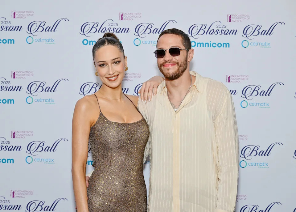 Two people posing on the red carpet at a celebrity event; one in a sparkling gown and the other in a casual shirt with sunglasses