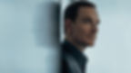 A man in a suit leans against a reflective wall, looking contemplative. Soft lighting and muted tones create a calm, introspective mood.