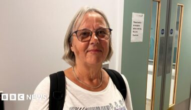 Renate Kennedy in A&E in Bournemouth. She has shoulder length blonde hair and is wearing a white top with the black straps of a bag over her shoulders. She is wearing glasses and a silver necklace. She is standing in front of a white wall with green double doors.
