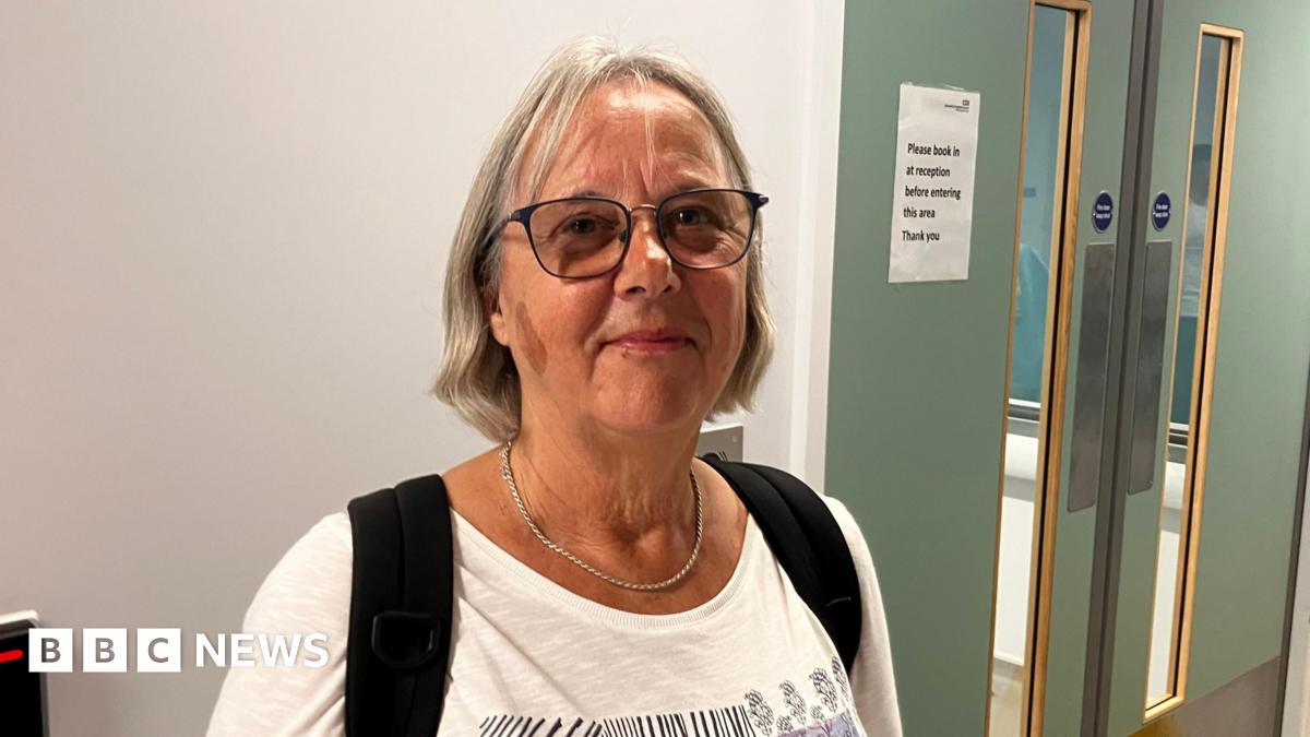 Renate Kennedy in A&E in Bournemouth. She has shoulder length blonde hair and is wearing a white top with the black straps of a bag over her shoulders. She is wearing glasses and a silver necklace. She is standing in front of a white wall with green double doors.