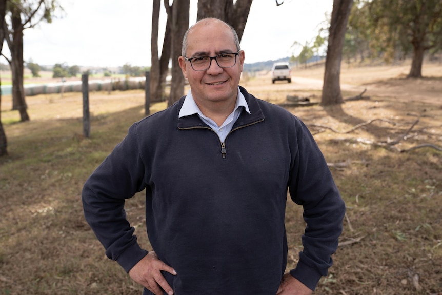 A man wearing glasses and a jumper stands under trees.