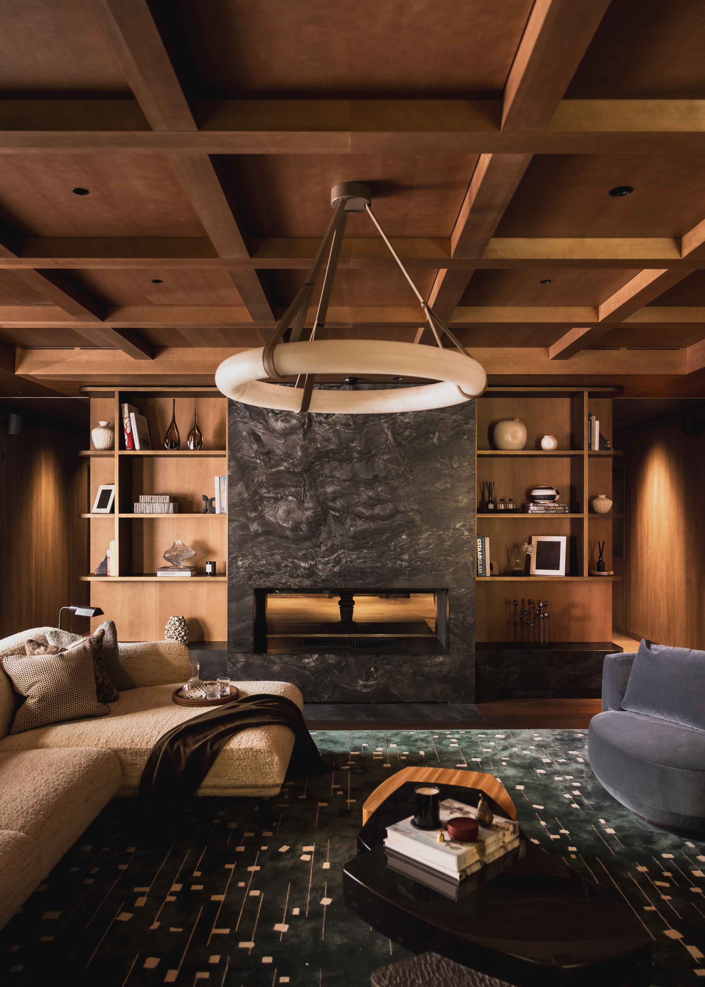 Snug with wood-paneled walls and ceiling, dark grey-green rug, beige chaise longue sofa and grey marble fireplace
