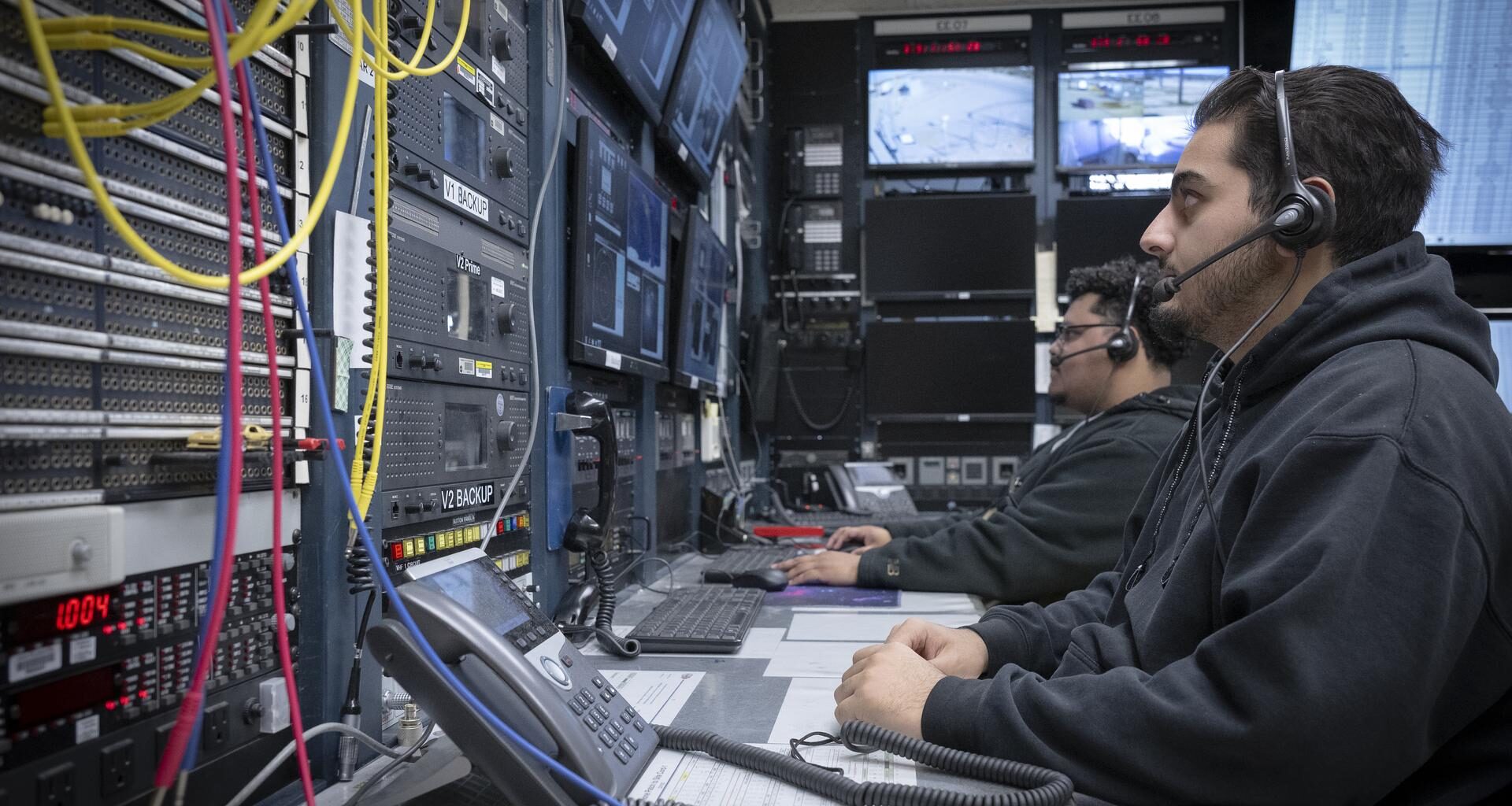 Two men wearing headsets sit in front of a wall of screens and instrumentation.