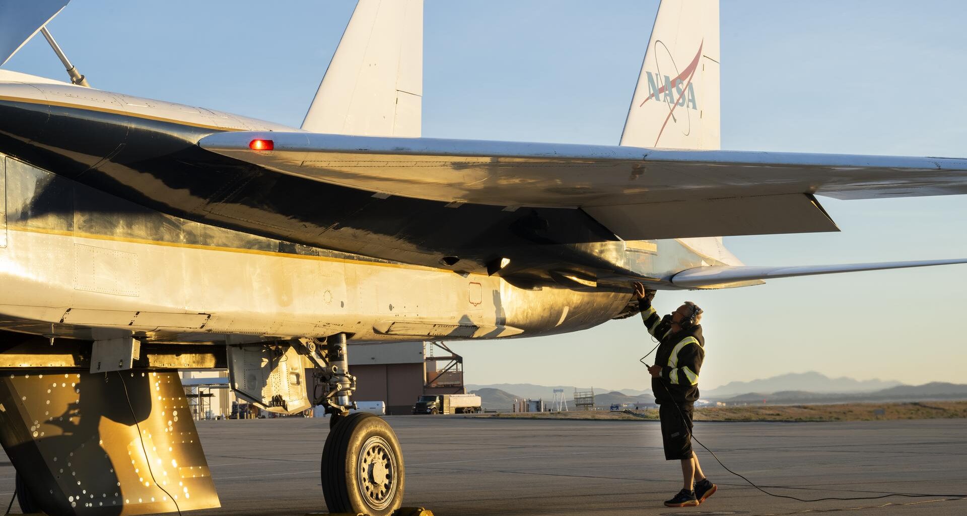 The back of a white jet aircraft’s wing and tails are seen in this image. One wheel and an attached test object, shaped like a wing, hangs from the bottom of the jet. A man in a black jacket, with yellow reflective coating, stands underneath and raises one hand to the aircraft. The man is wearing ear protection. Desert sand and mountains are seen in the background.