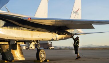 The back of a white jet aircraft’s wing and tails are seen in this image. One wheel and an attached test object, shaped like a wing, hangs from the bottom of the jet. A man in a black jacket, with yellow reflective coating, stands underneath and raises one hand to the aircraft. The man is wearing ear protection. Desert sand and mountains are seen in the background.