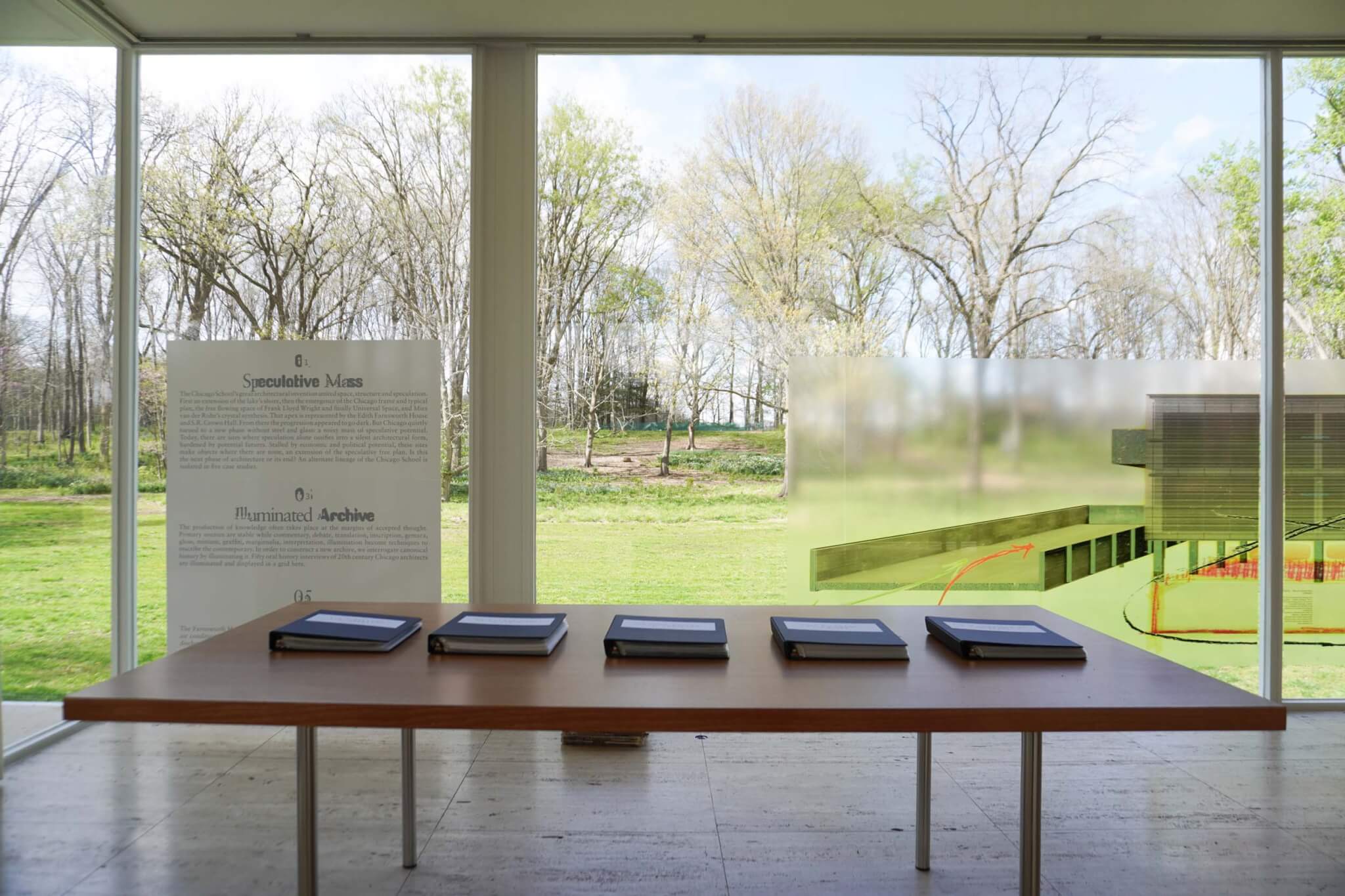 Five dark blue binders arranged in a row on a wooden dining table inside the glass-walled Edith Farnsworth House