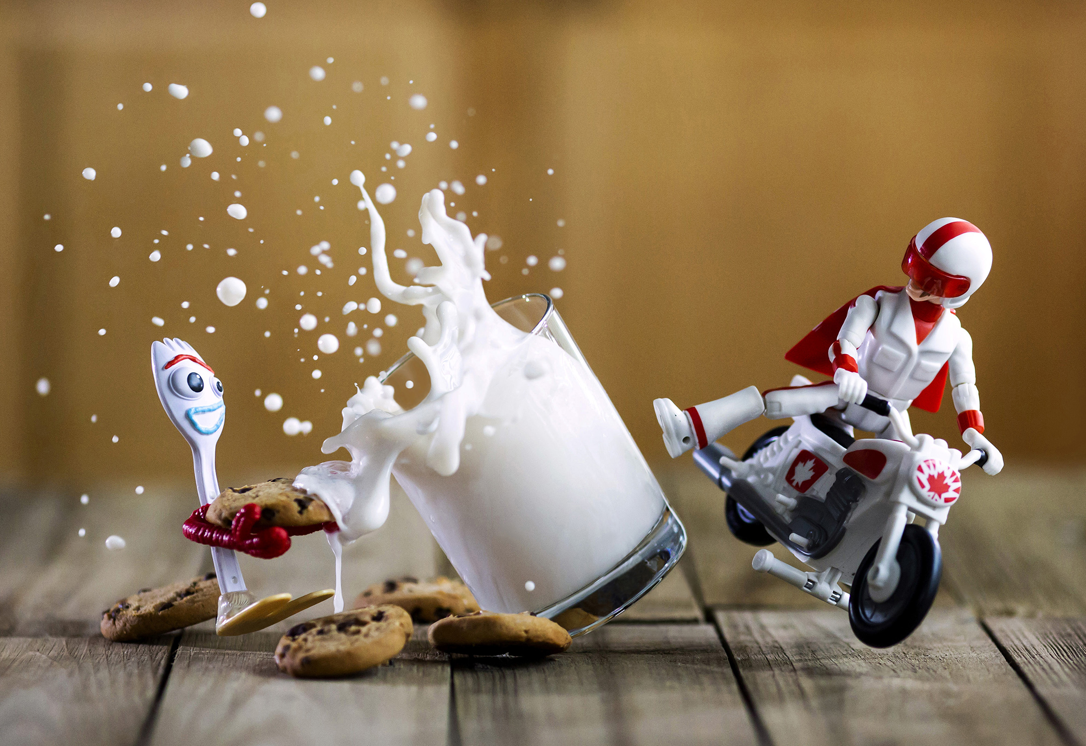 Toy fork holding cookies splashes milk from a tipping glass as a toy motorcyclist in red and white performs a stunt nearby