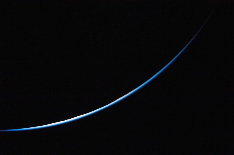 A thin, glowing blue curve of Earth's atmosphere is seen on the dark edge of the planet, set against the blackness of space, creating a crescent-like shape.