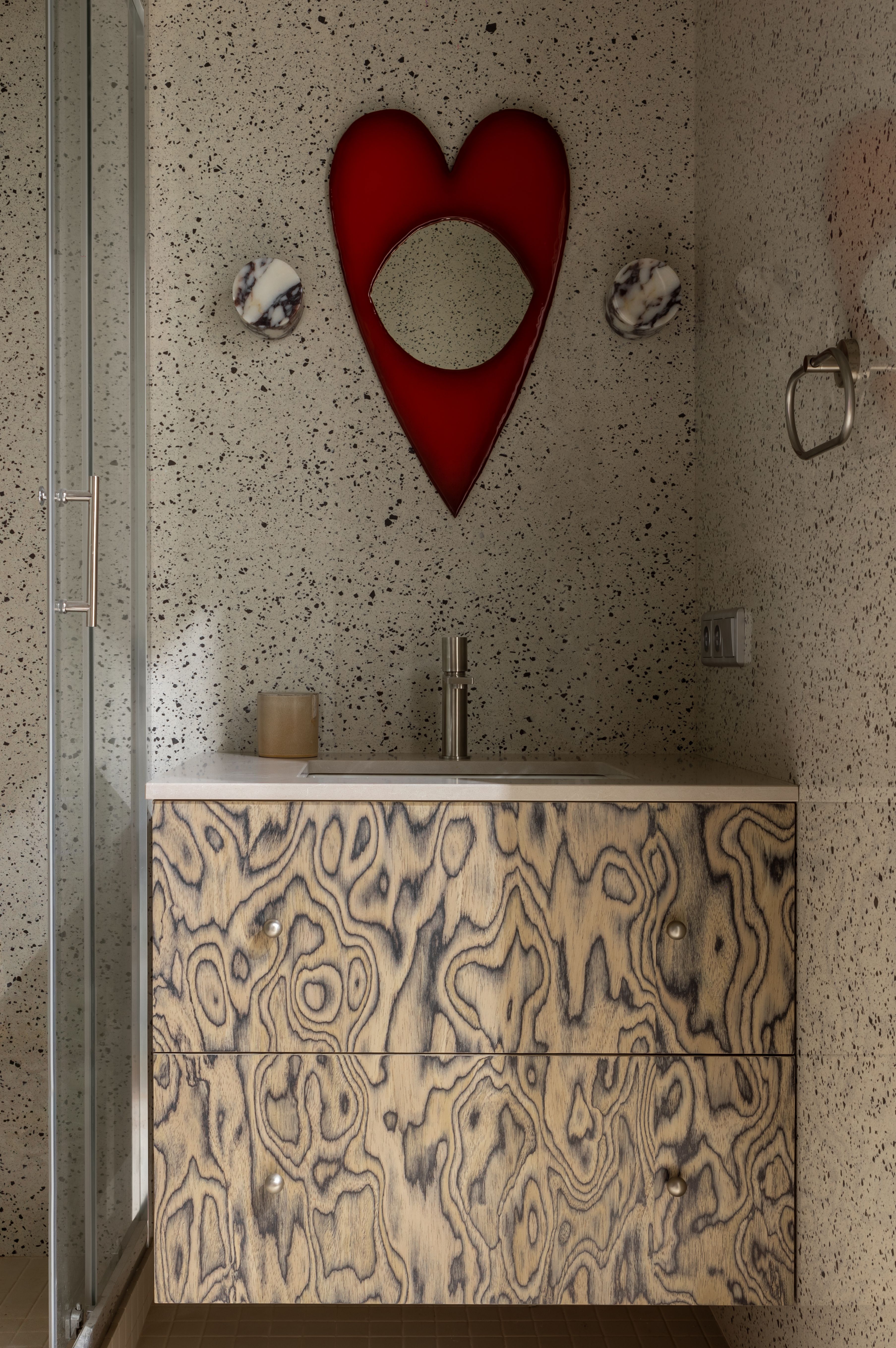 A bathroom with terrazzo walls, wood veneer vanity, and heart-shaped mirror 