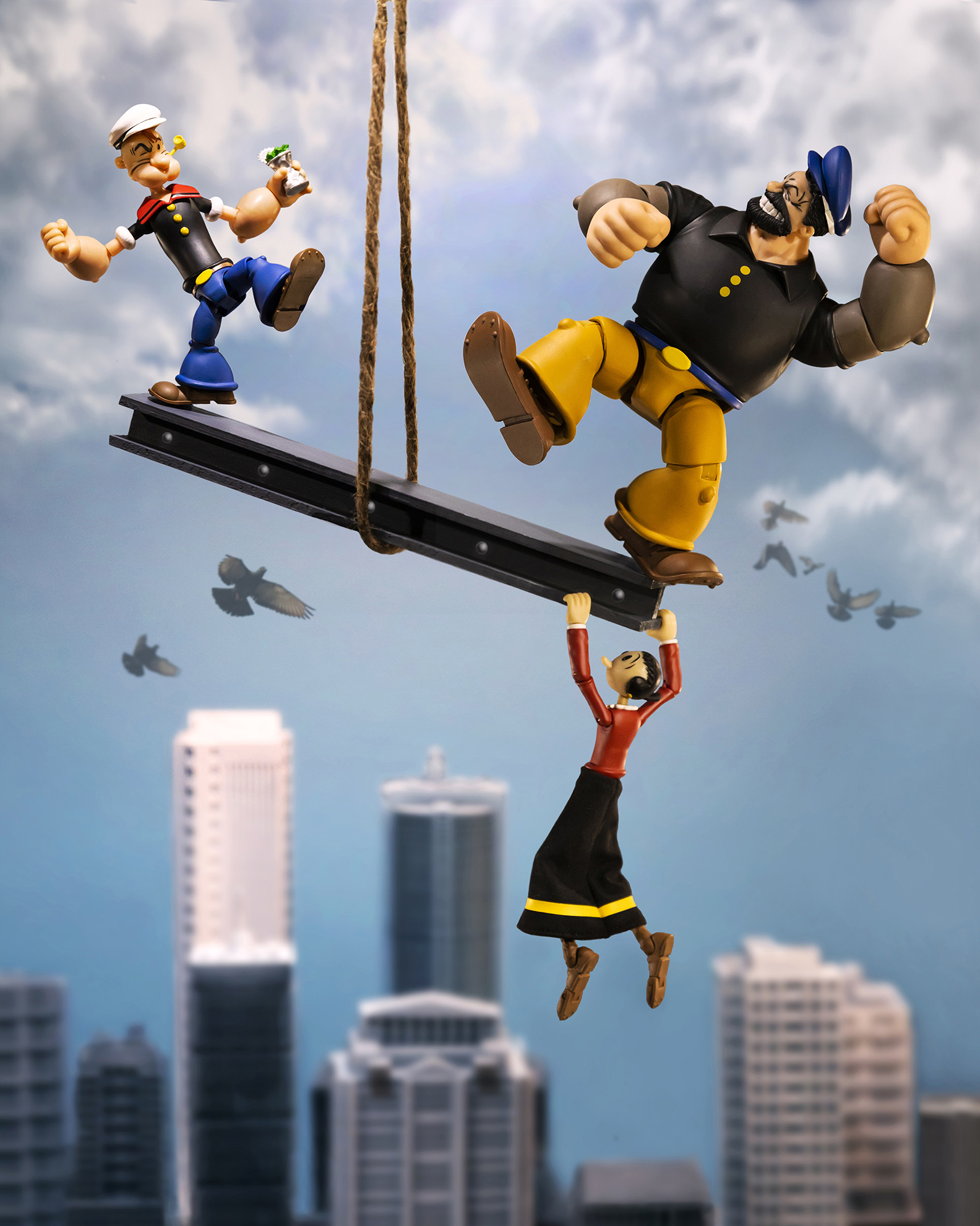 Action figures of Popeye characters on a suspended beam, with Olive Oyl hanging below and city buildings in the background