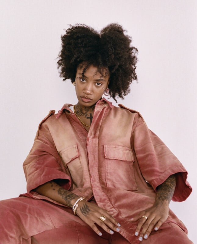A person with natural curly hair sits against a light background, wearing an oversized dusty pink jacket and pants. They have visible tattoos, gold jewelry, and a calm, confident expression.