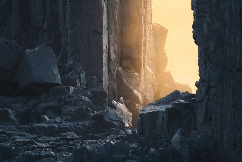 Sunlight streams through a narrow gap between tall, dark rock cliffs, illuminating jagged boulders and casting a golden glow on the rocky terrain below.