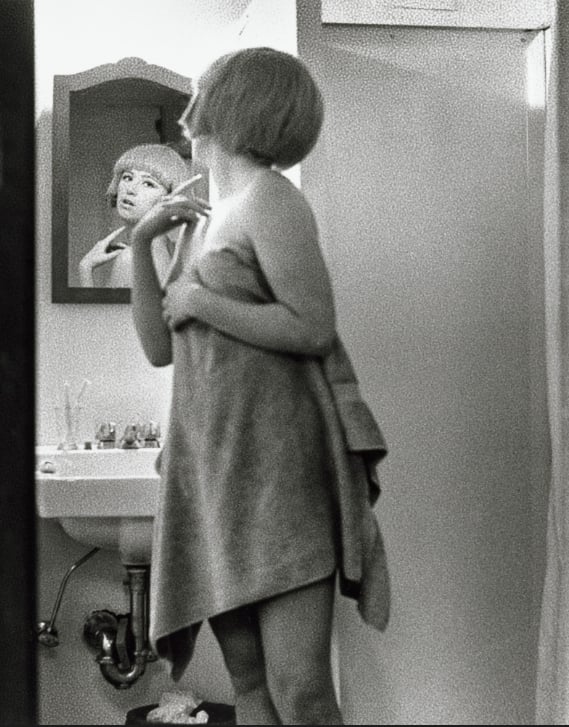 A person with short hair, wrapped in a towel, stands in a bathroom facing a sink. Their reflection is visible in the mirror, showing a thoughtful expression. The image is in black and white.