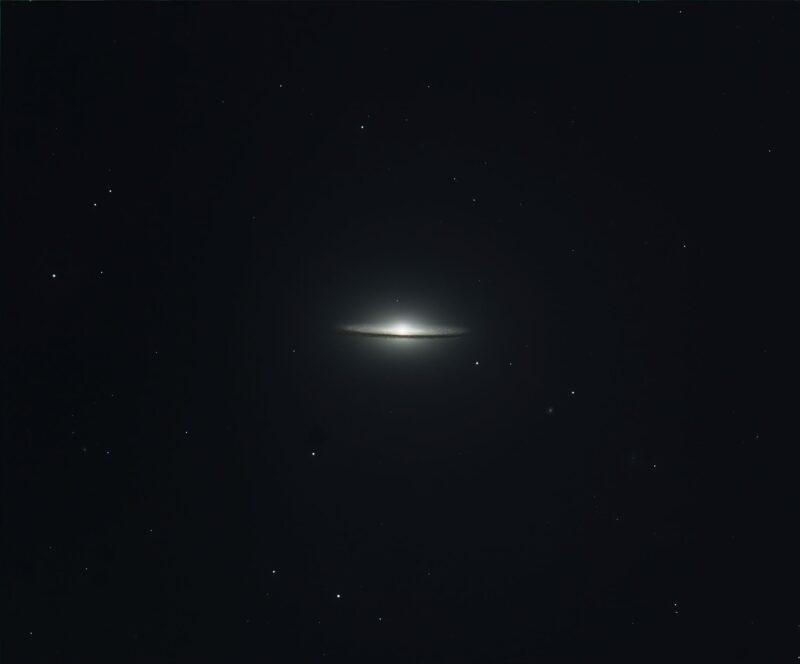 Whitish hat-shaped galaxy seen edge-on, with a prominent dark lane and sparse foreground stars.