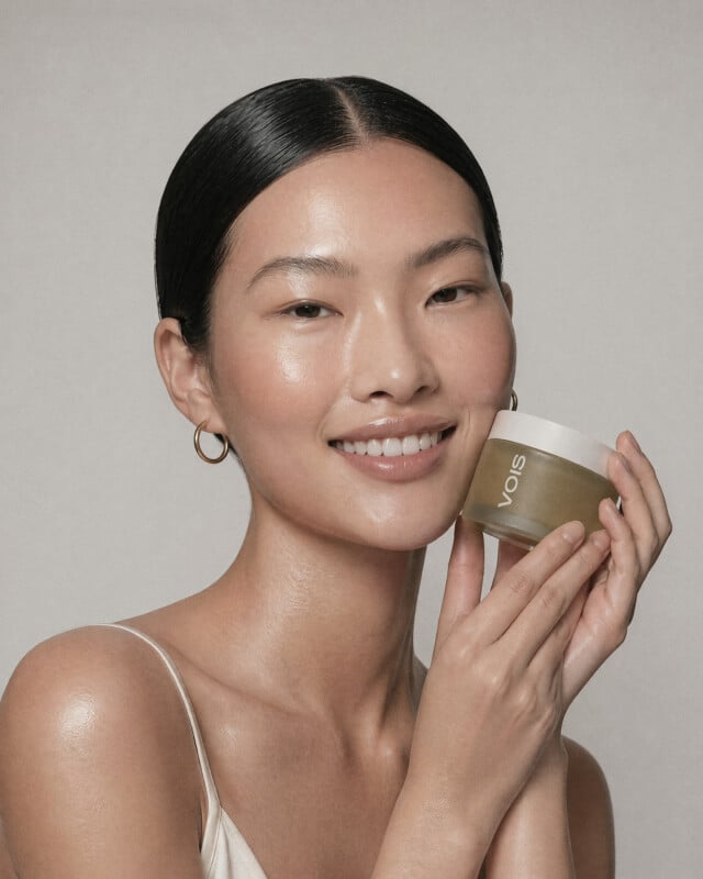 A woman with smooth, glowing skin smiles while holding a gold VOIS skincare jar near her face. She has straight black hair pulled back and wears small hoop earrings and a white tank top.