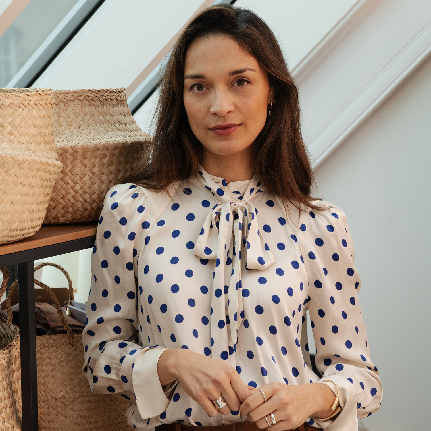 A woman with long brown hair in a blue and cream polka dot blouse by a shelf of woven baskets