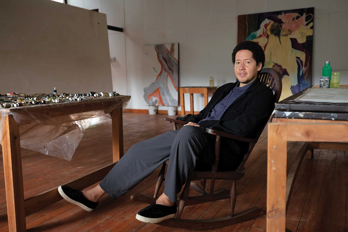 Person sitting in a rocking chair in an art studio with paintings and art supplies in the background.