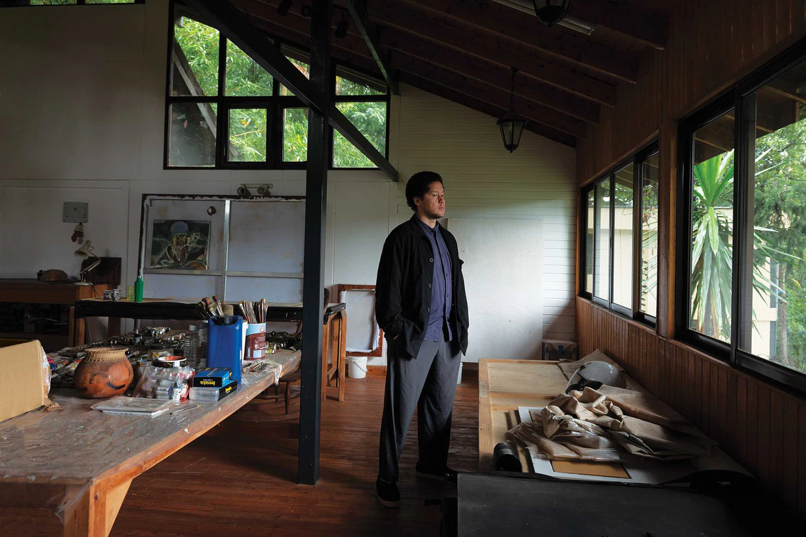 Person standing in a rustic studio with large windows, surrounded by art supplies and natural light.