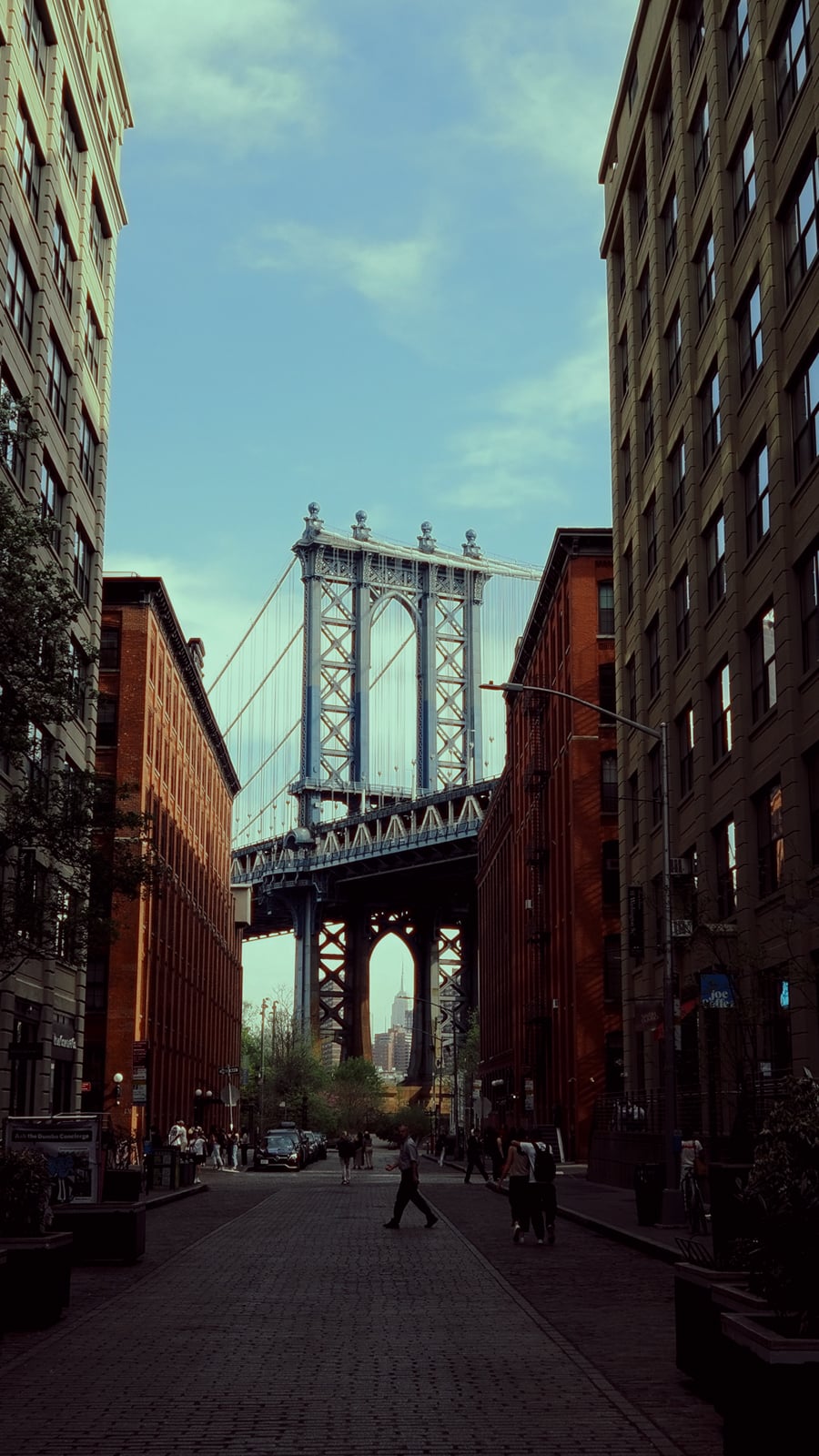 View of the Manhattan Bridge framed by tall brick buildings in Brooklyn, with the Empire State Building visible through the bridge’s arch on a partly cloudy day. Pedestrians and cars are seen on the cobblestone street.