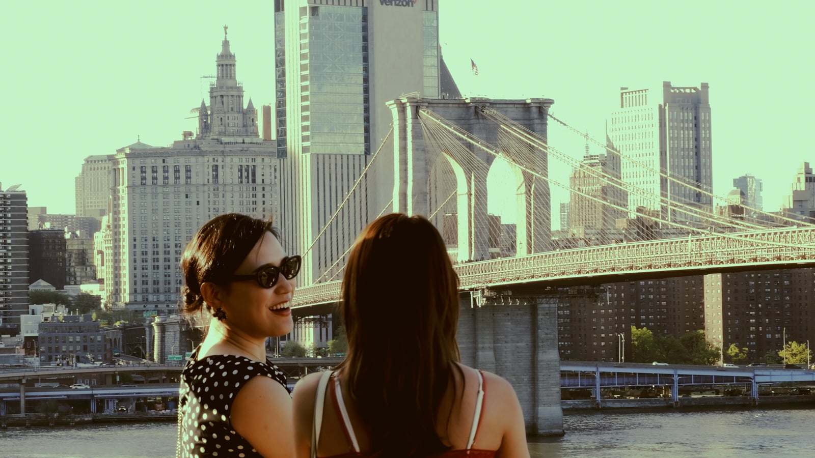 Two women stand by the water, one smiling and wearing sunglasses, with the Brooklyn Bridge and New York City skyline in the background on a sunny day.