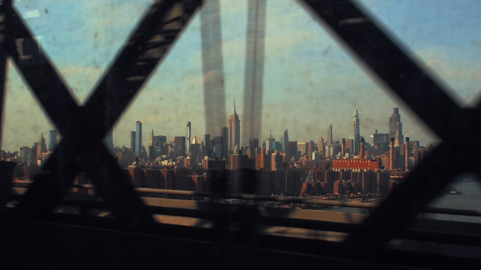 A view of the New York City skyline, including the Empire State Building, seen through the dark, crisscrossing beams of a bridge on a hazy day.