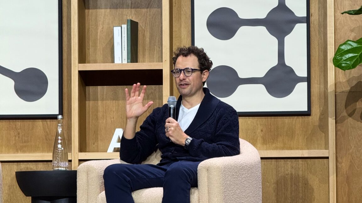 Dario Amodei sits on a chair on stage and gestures as he talks.