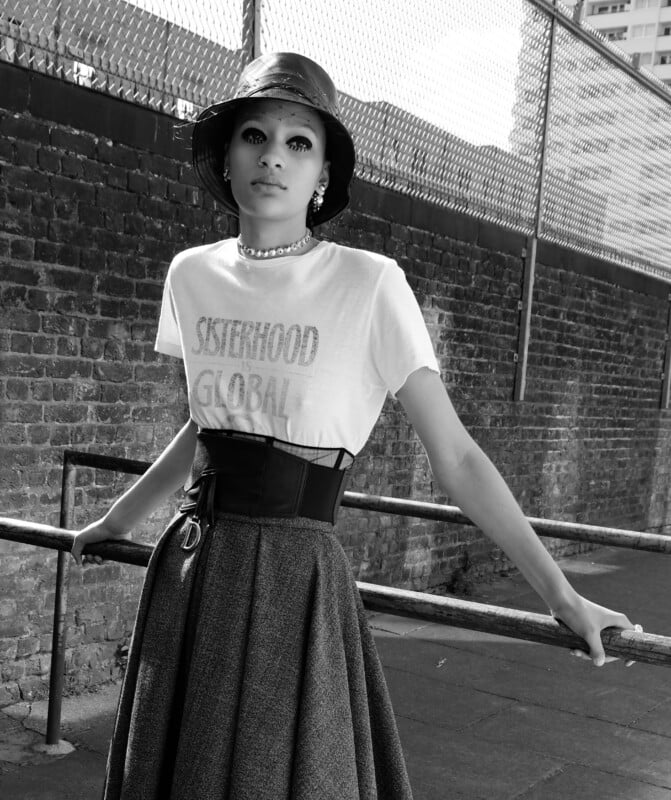 A stylish person in a bucket hat, choker, and "Sisterhood Global" t-shirt leans against a railing outdoors, wearing a high-waisted skirt and corset belt, with a brick wall and chain-link fence in the background.