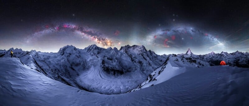 A panoramic view of snow-covered mountains under a starry night sky with the Milky Way visible; a person in yellow stands on the left, and a glowing orange tent sits on the right ridge.