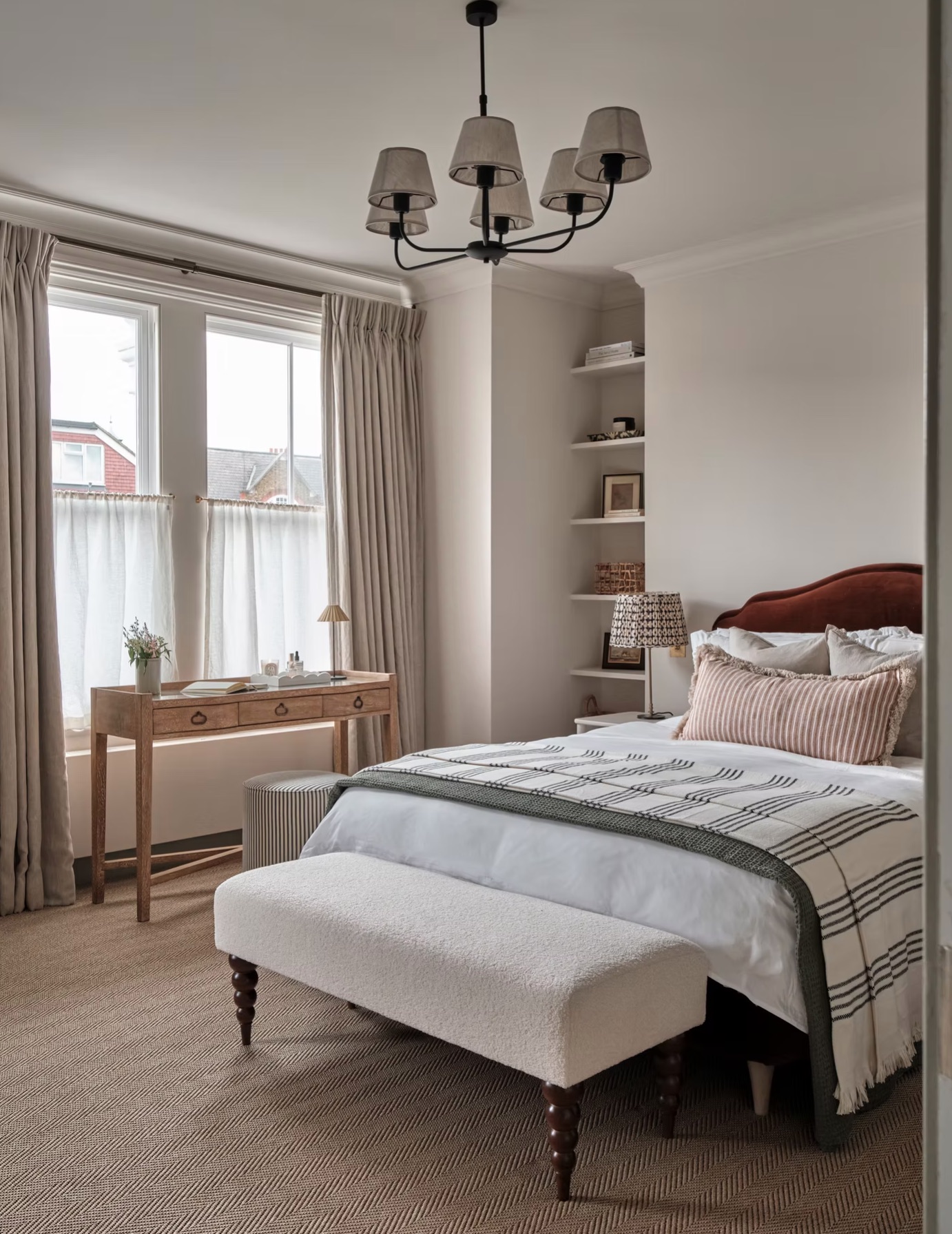 A bedroom with a wooden desk under a large window, shelves built into an alcove, and a bed with a rust-colored velvet headboard, a striped throw, and a rectangular boucle bench with walnut bobbin legs