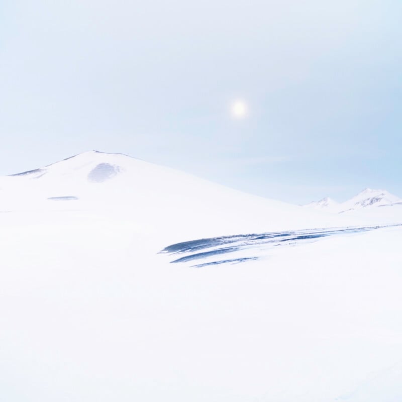 A bright, snowy landscape with gentle hills under a pale sky. The sun is faintly visible above the horizon, casting soft light over the white, wintery scene.
