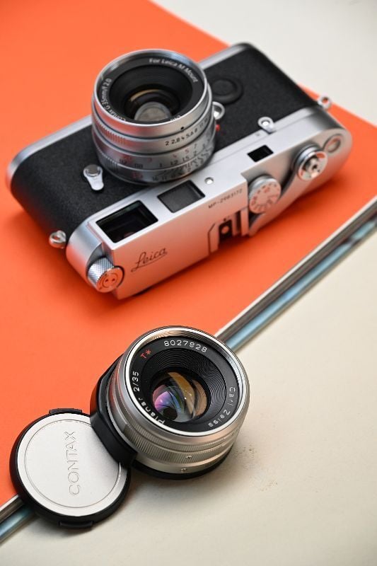 A silver Leica camera with a mounted lens rests on an orange folder, beside a silver Contax lens with its cap, all placed on a cream-colored surface.