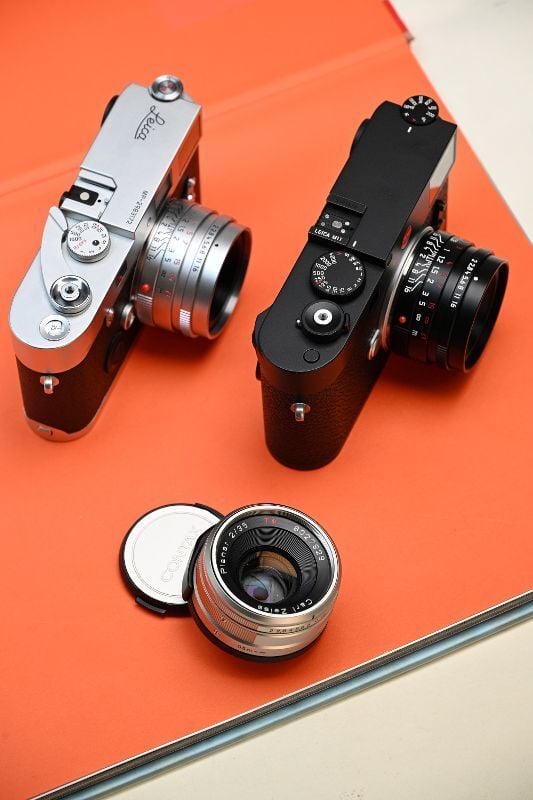 Two vintage cameras, one silver and one black, are displayed on an orange surface with a detached camera lens and its cap placed nearby.
