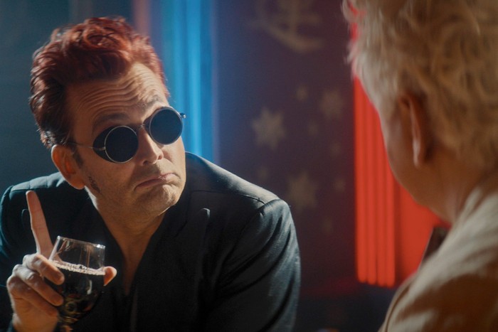 David Tennant as Crowley in Good Omens season 3, with red hair and black sunglasses, holding a glass and looking at Michael Sheen's Aziraphale.