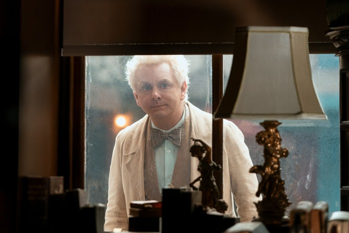 Michael Sheen as Aziraphale in Good Omens season 3, clad in white and peering through a window in