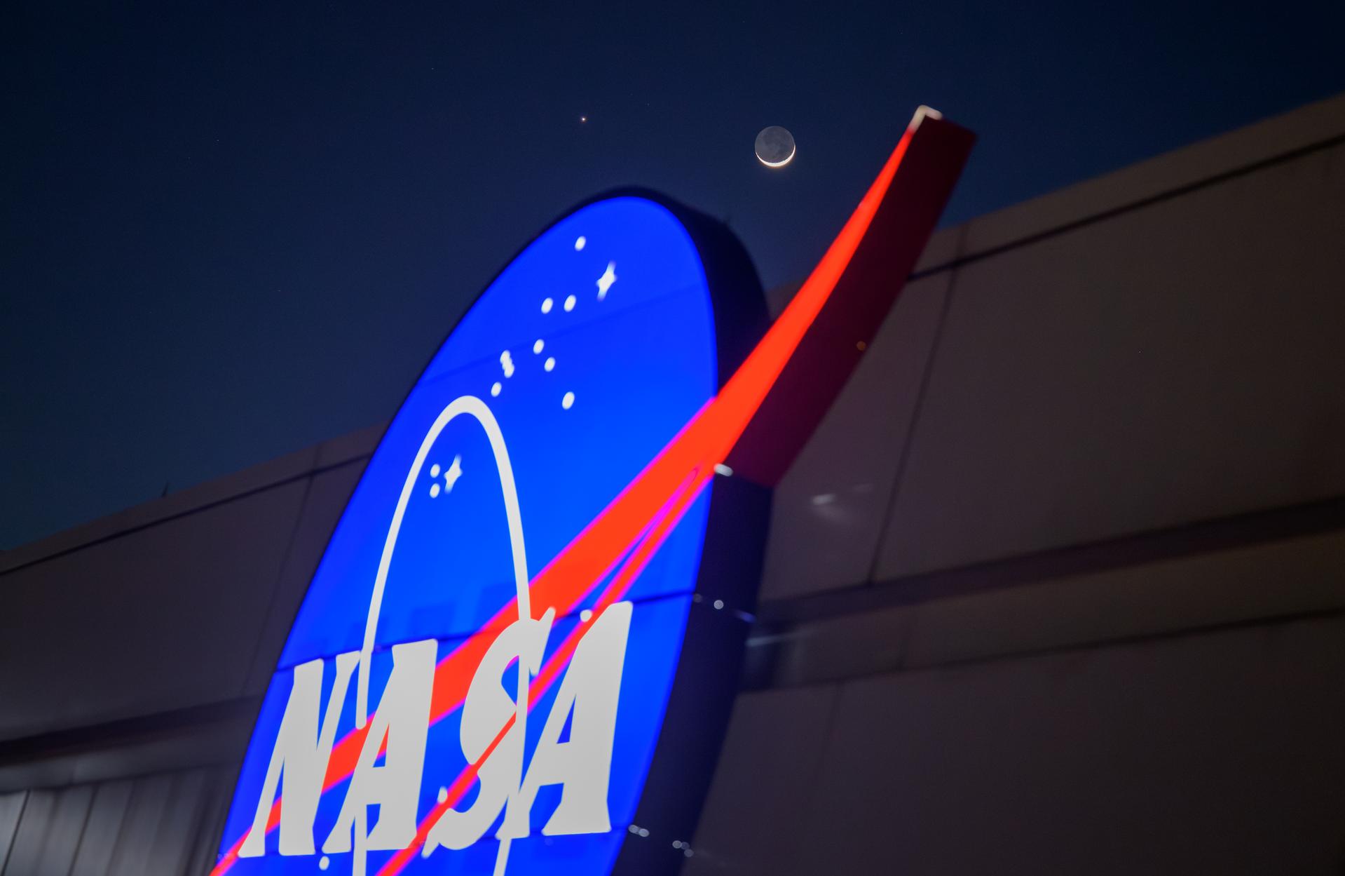 The crescent moon and Venus shine in the night sky above the NASA "Meatball" logo on the side of a building.